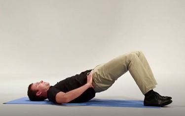 A White man is lying on his back on a mat with his hips elevated, knees bent, and feet flat on the ground.