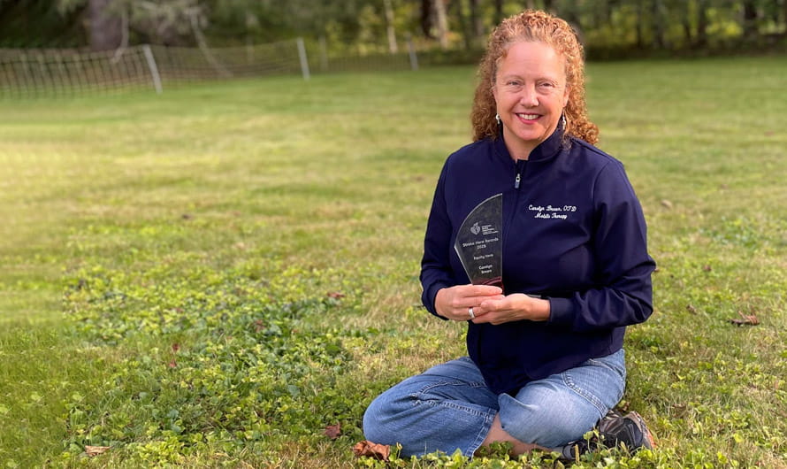 Carolyn Brown is sitting outside in the grass holding her Stroke Hero Award trophy.