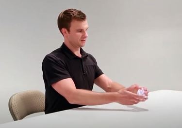 A White man is sitting at a table with his arms outstretched in front of him. His hands are holding a crumpled ball of paper.