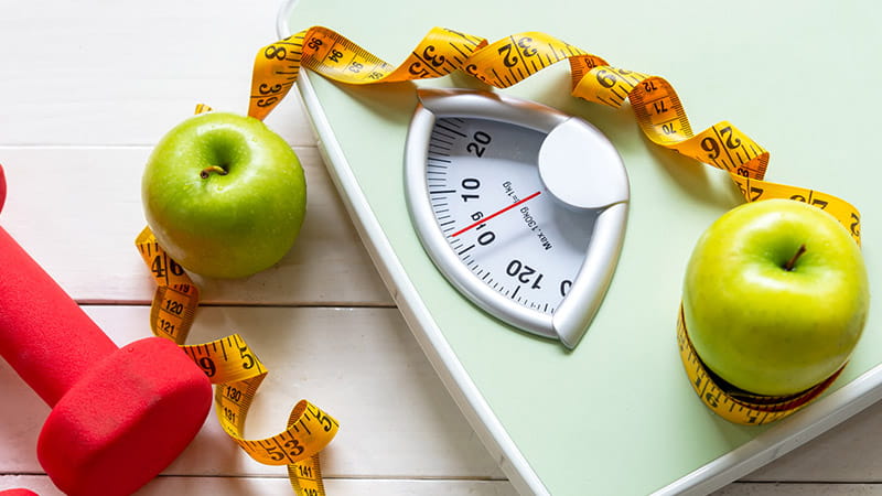 Green apples, a red dumbbell and measuring tape are arranged around a body scale.