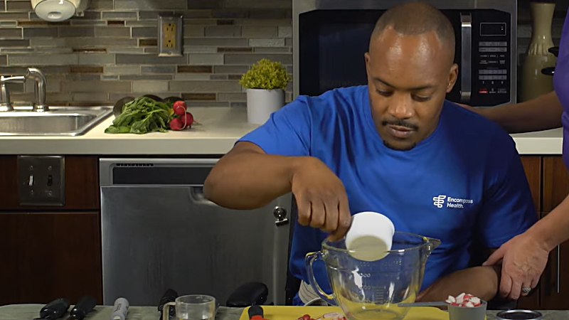 A man in a wheelchair is pouring food from a measuring cup into a glass bowl.