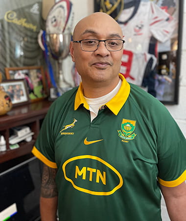 Stroke survivor, Silivenusi “Nusi” Tukuafu, is standing in front of sports memorabilia wearing a Springboks rugby jersey.