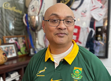 Stroke survivor, Silivenusi “Nusi” Tukuafu, is standing in front of sports memorabilia wearing a Springboks rugby jersey. Stroke survivor, Silivenusi “Nusi” Tukuafu, is standing in front of sports memorabilia wearing a Springboks rugby jersey.