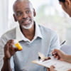 A middle-aged Black man is showing a bottle of prescription medication to a female medical professional taking notes on a clipboard.
