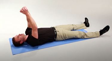 A White man is lying on his back on a mat with his arms extended straight up and hands clasped.