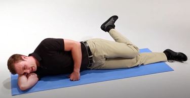 A White man is lying on his side with one arm supporting his head. His left knee is bent, and his left foot is extended behind him.
