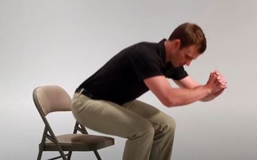 A White man is in a sitting position hovering over the seat of a chair. His hands are clasped in front of him for balance.
