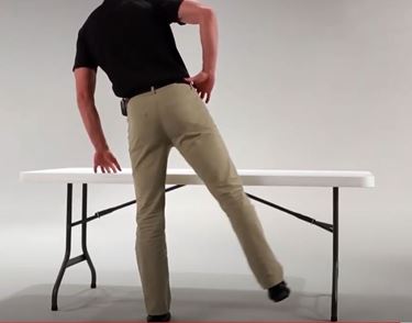 A White man is leaning on his left leg with his right leg out to his side. His left hand is resting on a folding table for support.