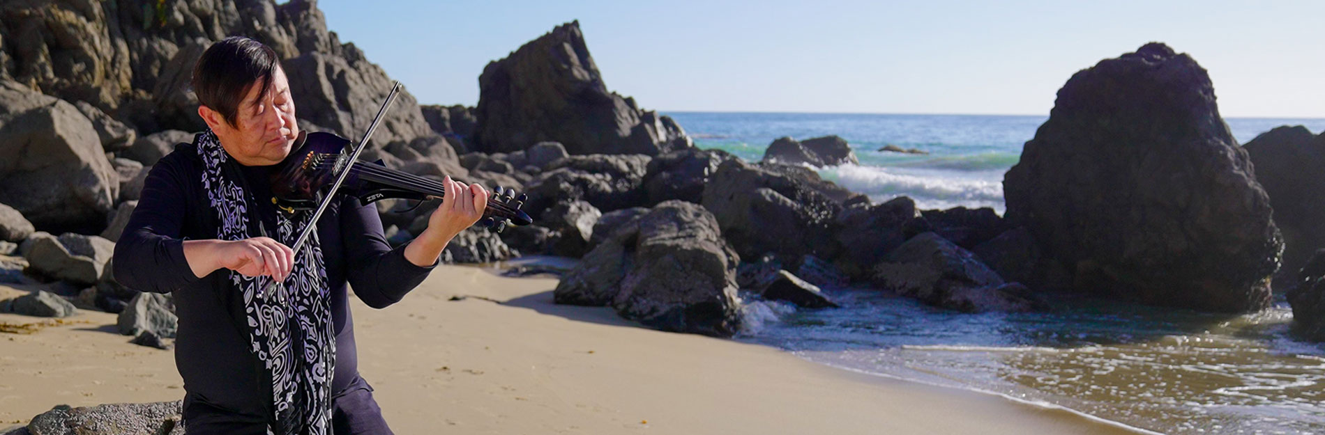 Stroke survivor, Dave, is playing a violin on the beach with the ocean and rocks in the background.