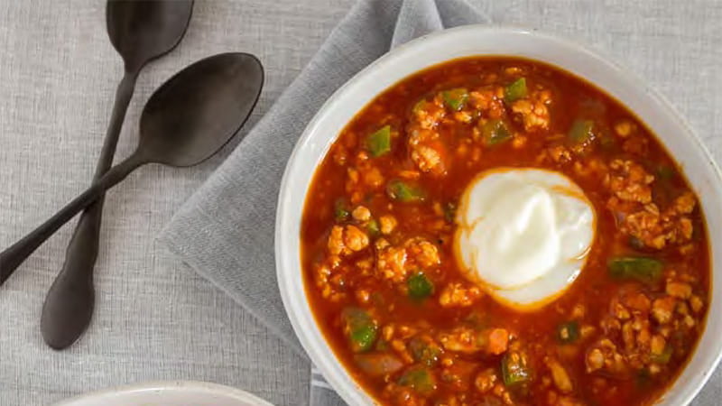 an aerial view of a bowl of chili with a dollop of yogurt and spoons arranged on grey table linens