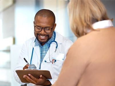 A smiling Black doctor is taking notes while talking with his senior female patient.