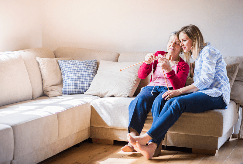 An older adult woman is sitting on a sofa knitting with a young adult woman. An older adult woman is sitting on a sofa knitting with a young adult woman.