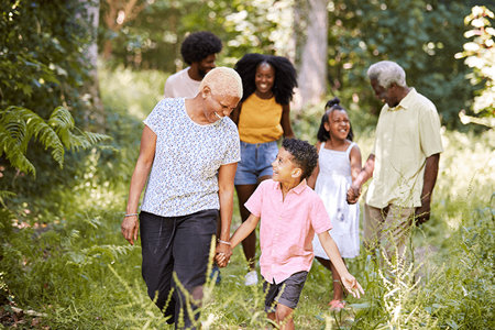 A senior Black woman and man are walking with their multigenerational family along a nature trail through trees.